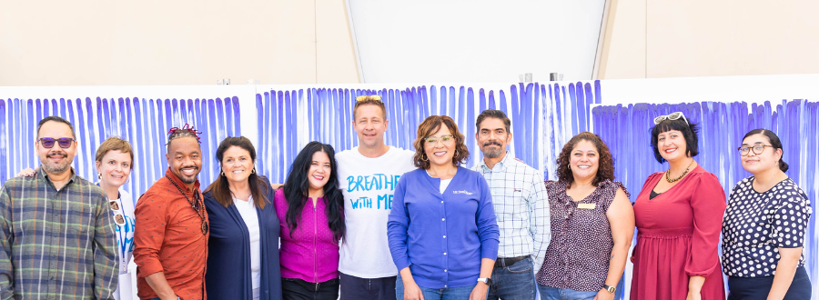 7 of 8, Group of adult staff with Jeppe Hein, a white man in a white shirt that says "Breathe with Me" stand in front of a white canvas covered with blue lines. 